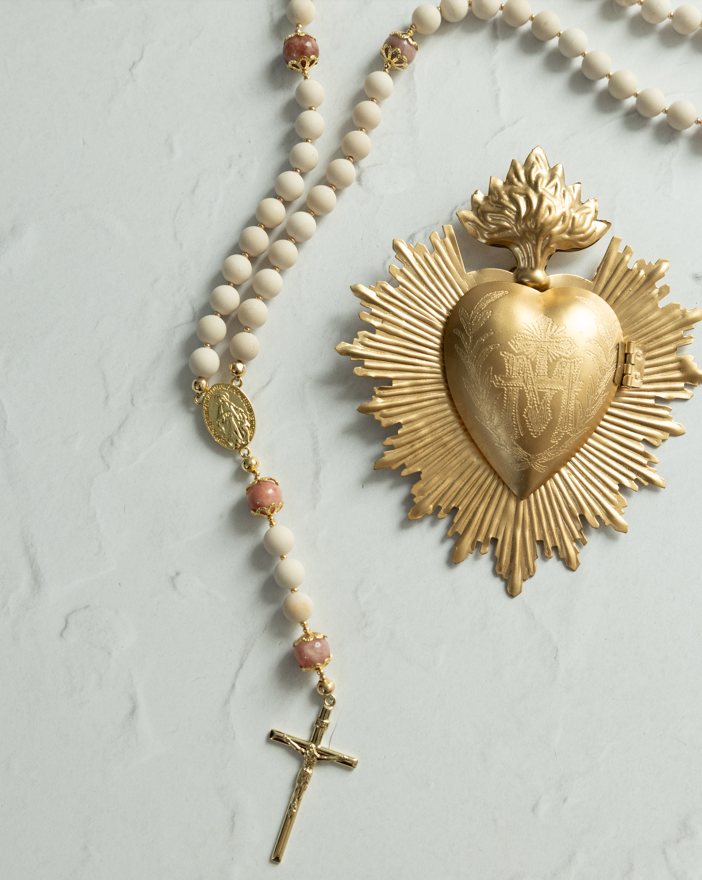 Golden sacred heart medal with sunburst design and engraved details, accented by a cream-colored beaded rosary featuring pink gemstone beads.