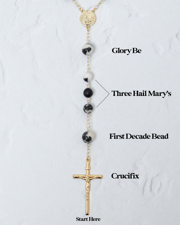Gold-toned rosary with black and white speckled beads, a gold crucifix, and a circular medallion at the top.
