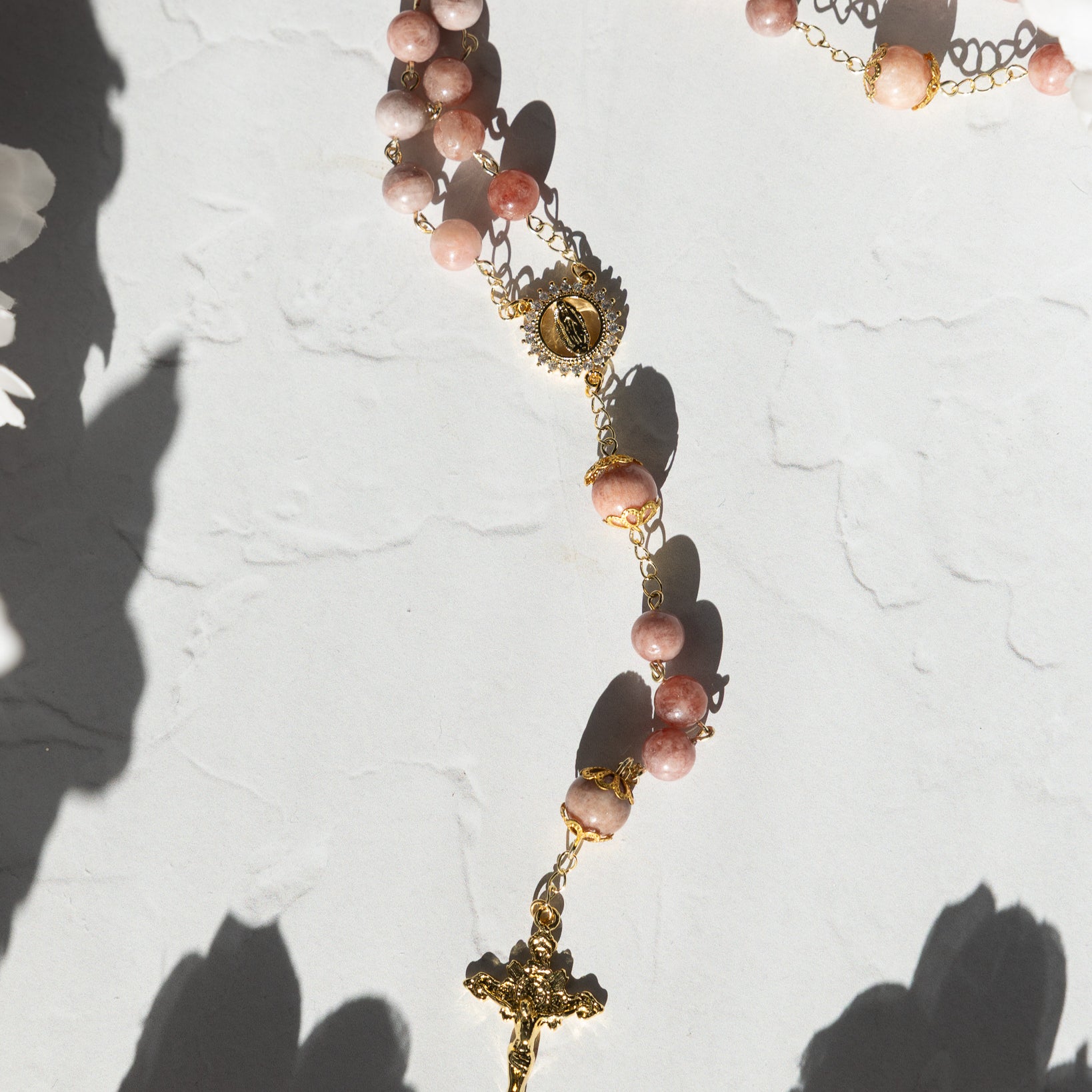 A delicate rosary featuring soft pink, translucent gemstone beads strung on gold-toned metal chains with a detailed golden crucifix and an ornate medallion.