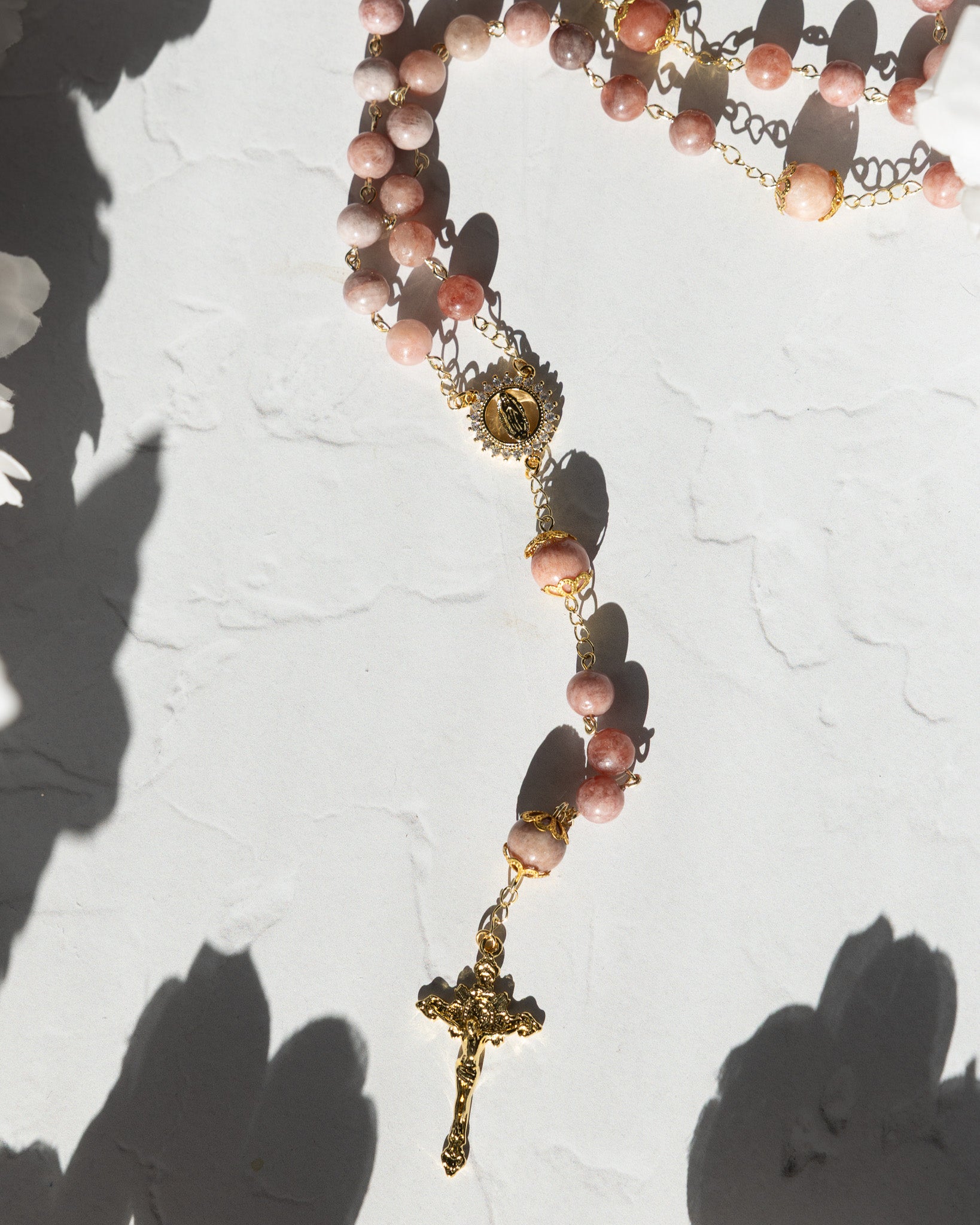 A delicate rosary featuring soft pink, translucent gemstone beads strung on gold-toned metal chains with a detailed golden crucifix and an ornate medallion.
