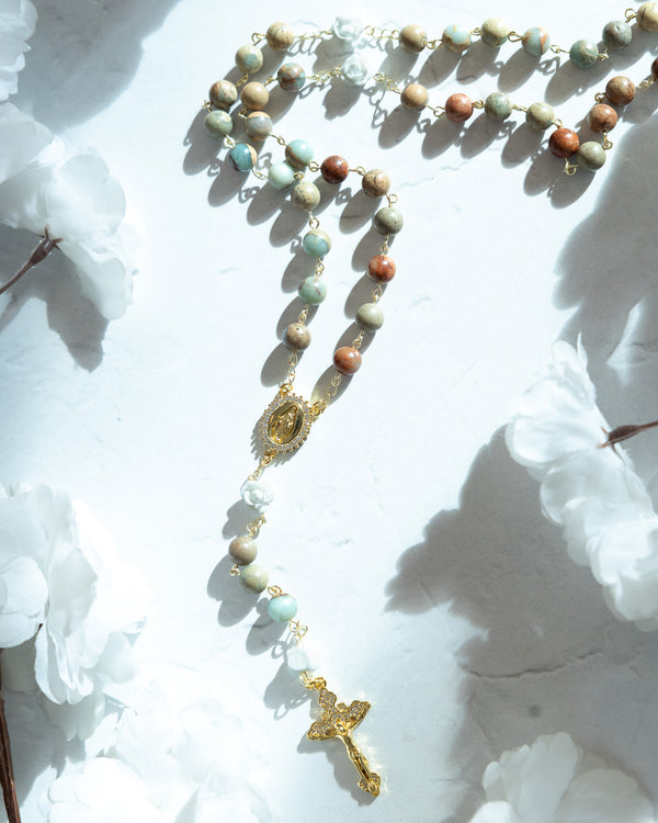 A delicate rosary featuring multicolored, earthy-toned beads in shades of green, beige, and brown with a gold-tone metal centerpiece medallion and cross.