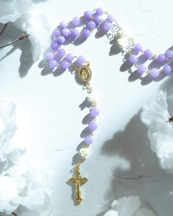A delicate lavender beaded rosary with gold-toned metal accents and a central religious medallion, accented by small white floral beads.