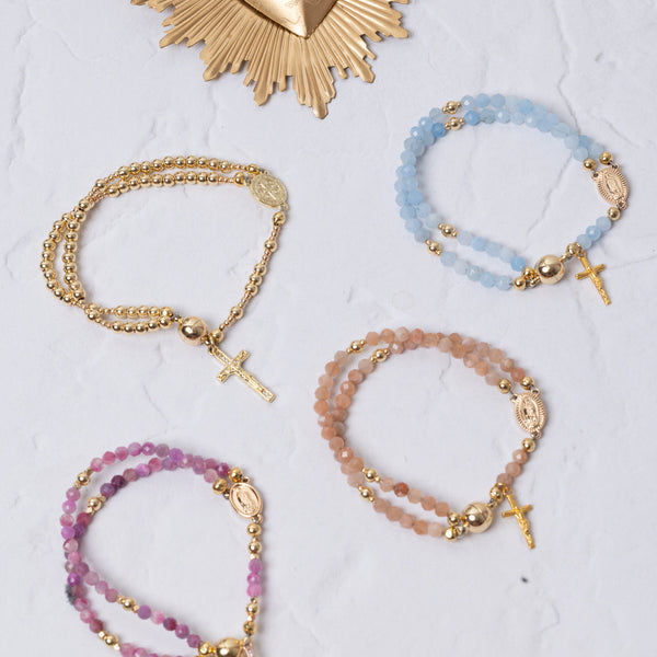 Set of four beaded rosary-style bracelets in pastel blue, pink, and peach tones with gold-toned metal accents and small crosses.