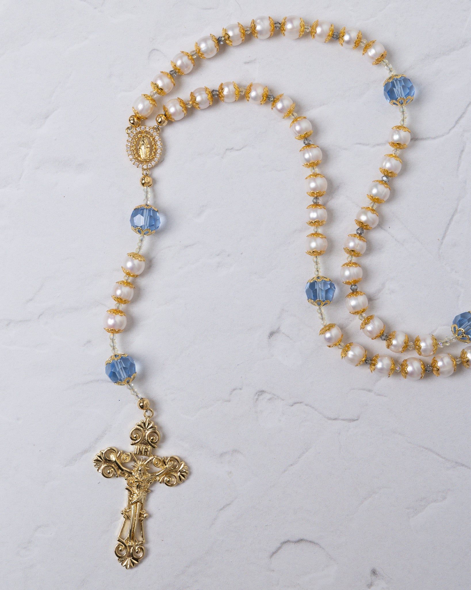 Elegant gold-toned rosary featuring alternating white pearls and faceted blue crystals, with an ornate crucifix pendant.