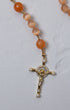 A gold-toned crucifix pendant with clear crystals and pearl accents on an orange carnelian bead poppy rosary
