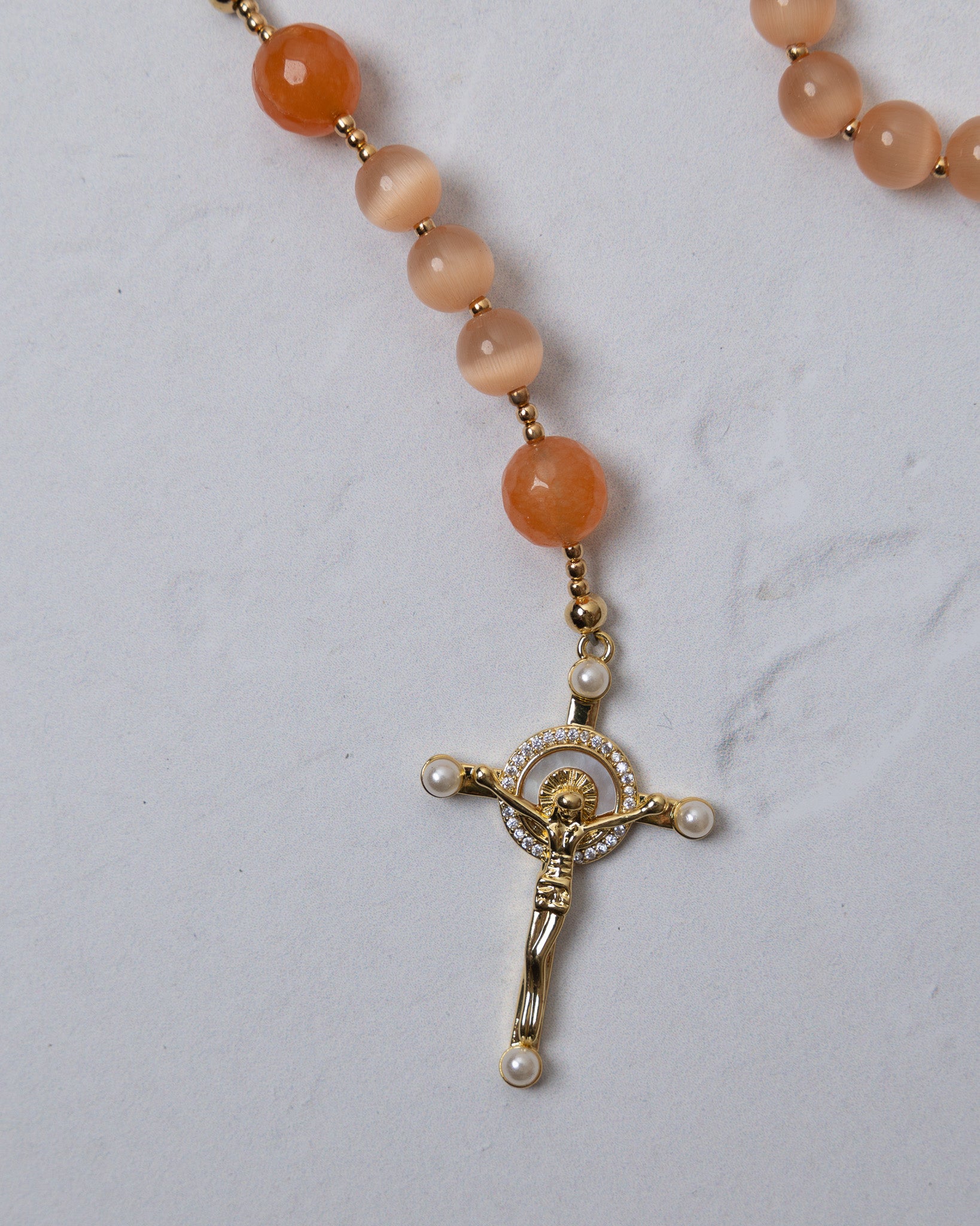 A gold-toned crucifix pendant with clear crystals and pearl accents on an orange carnelian bead poppy rosary