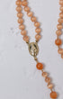 A delicate peach-colored cat eye gemstone rosary with gold-toned oval medallion featuring engraved religious figure