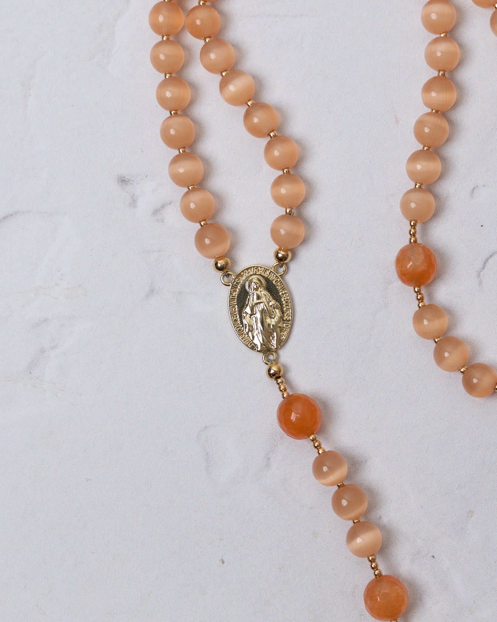 A delicate peach-colored cat eye gemstone rosary with gold-toned oval medallion featuring engraved religious figure