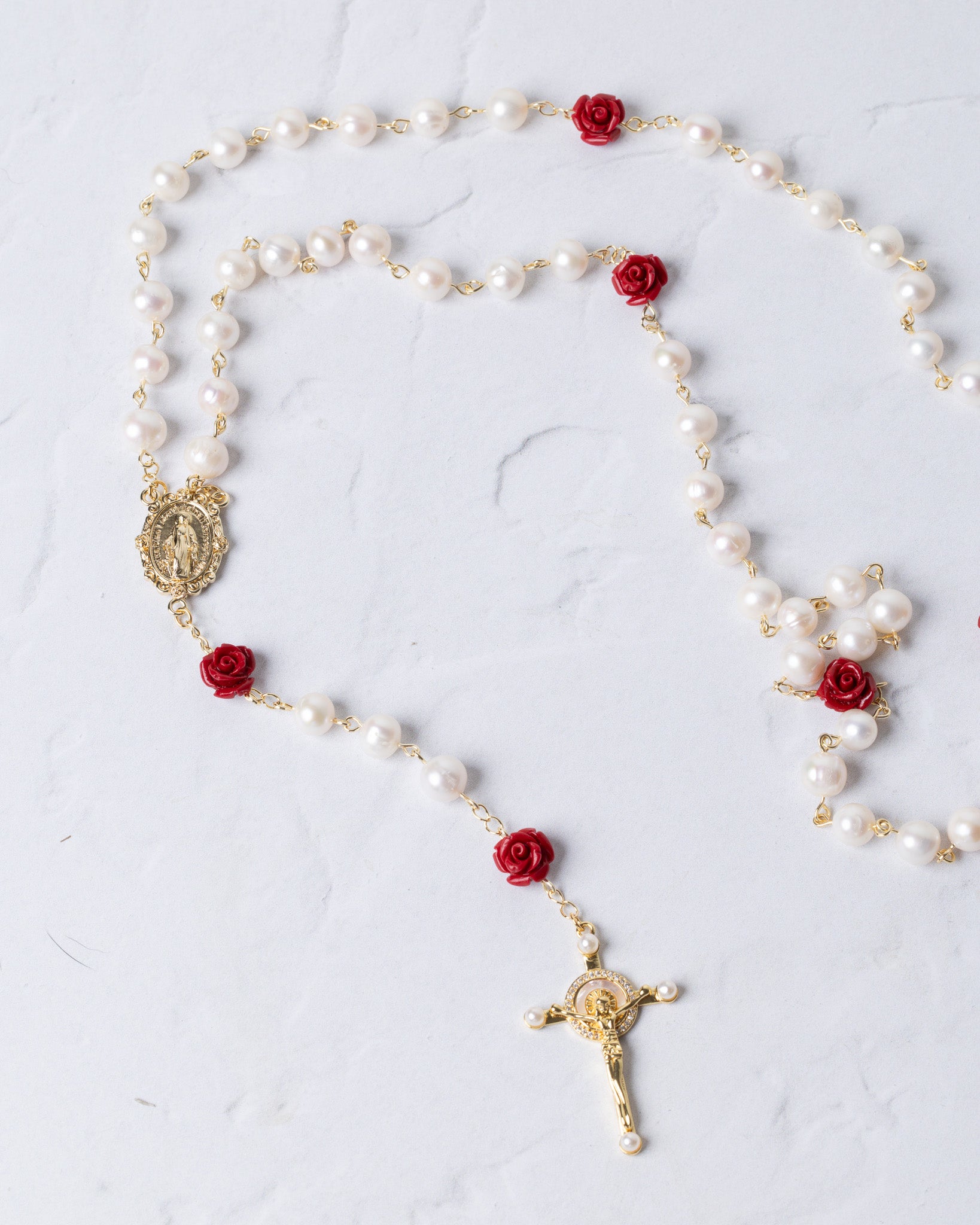 A delicate rosary featuring white pearls, red rose accents, and gold-toned metal components including a crucifix and an oval medallion.