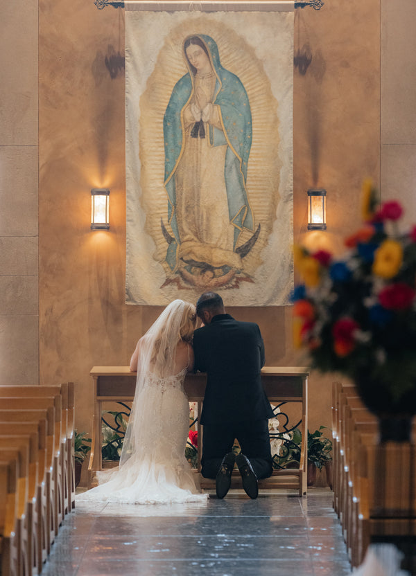 A bride in a flowing white wedding gown with lace detailing and a long veil kneels beside her groom, who is dressed in a black suit.