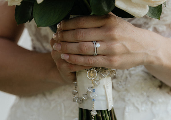 A delicate white rose bouquet held in hands, wrapped with a cream-colored ribbon and adorned with an ornate silver rosary featuring clear crystals.