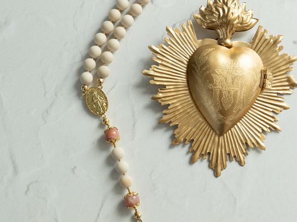 Golden sacred heart medal with sunburst design and engraved details, accented by a cream-colored beaded rosary featuring pink gemstone beads.